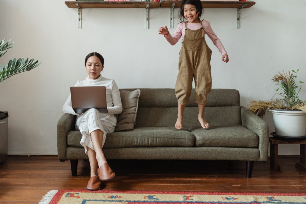 mother working from home while child jumps on couch 